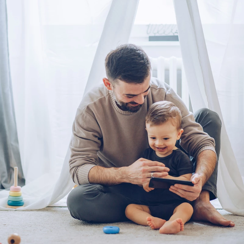 A man and a baby sit on the floor, engaged in play with a tablet, sharing a joyful moment together