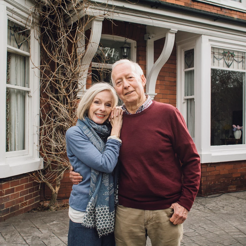 An older couple stands together in front of their house, smiling and enjoying a sunny day